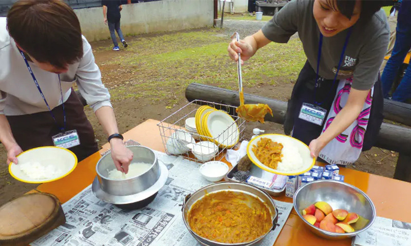 写真：レクリエーションの様子。左側の生徒はお釜からお皿へご飯を盛りつけ、右側の生徒は鍋からご飯の乗ったお皿へカレーを盛り付けている。