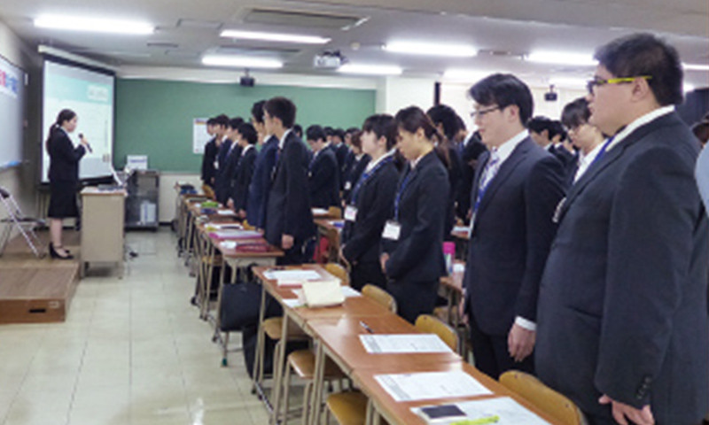 写真：就職集中講座受講の様子。学生たちは机の後ろに立ち、教壇の講師の話を聞いている。