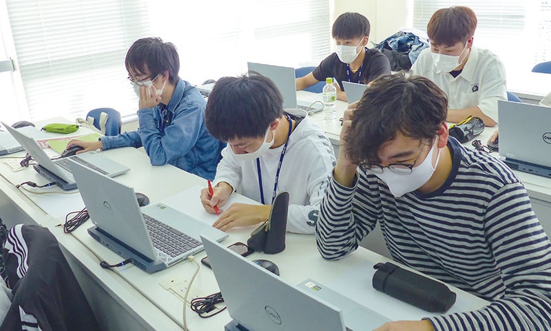 写真：プログラミングバトルの様子。学生5人がノートパソコンへ向かい作業をしている。