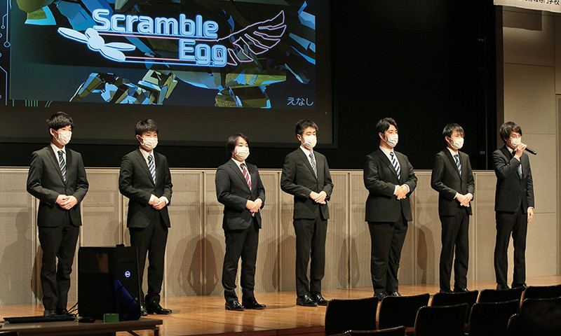 写真：卒業研究発表会の様子。7人の学生が壇上に立ち、作品「Scramble Egg」に付いて発表している。