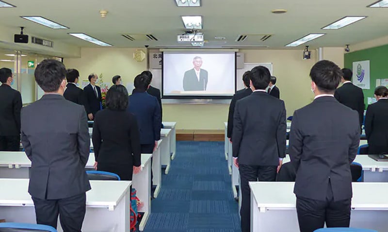 写真：北海道情報大学 学位記授与式の様子。参加者が正面のスクリーンに投影された登壇者の話を聞いている。