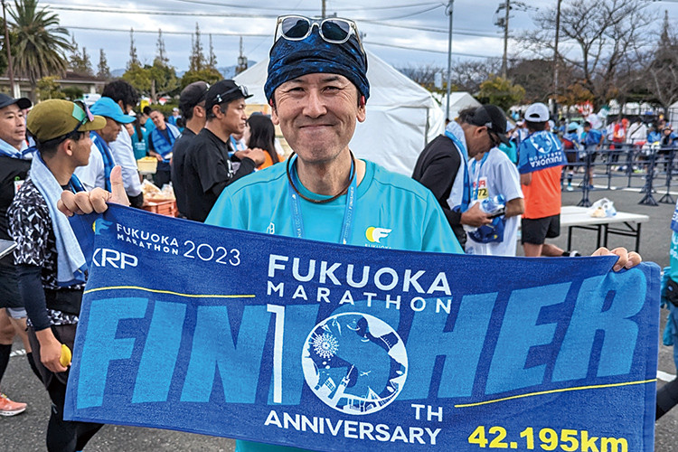 写真：正面を向き、笑顔で福岡マラソンのFINISHERのタオルを広げている大宮 貴昭先生。後ろにはたくさんの福岡マラソンの参加者が見える。