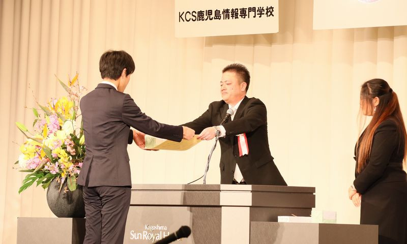 写真：KCS鹿児島情報専門学校の卒業式の様子。学生が校長から卒業証書を受け取っている。