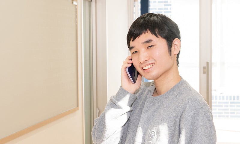 写真：グレーのスウェットを着た男子学生。スマートフォンで電話をかけている。