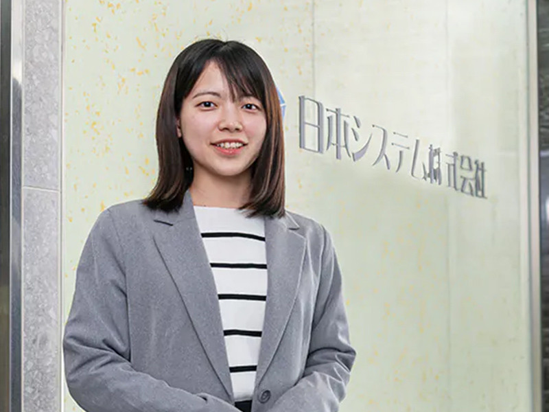写真：白と黒のボーターの服にグレーのジャケットを着た微笑んでいる女性。後ろの壁に日本システム株式会社のロゴマークが掲げられている。