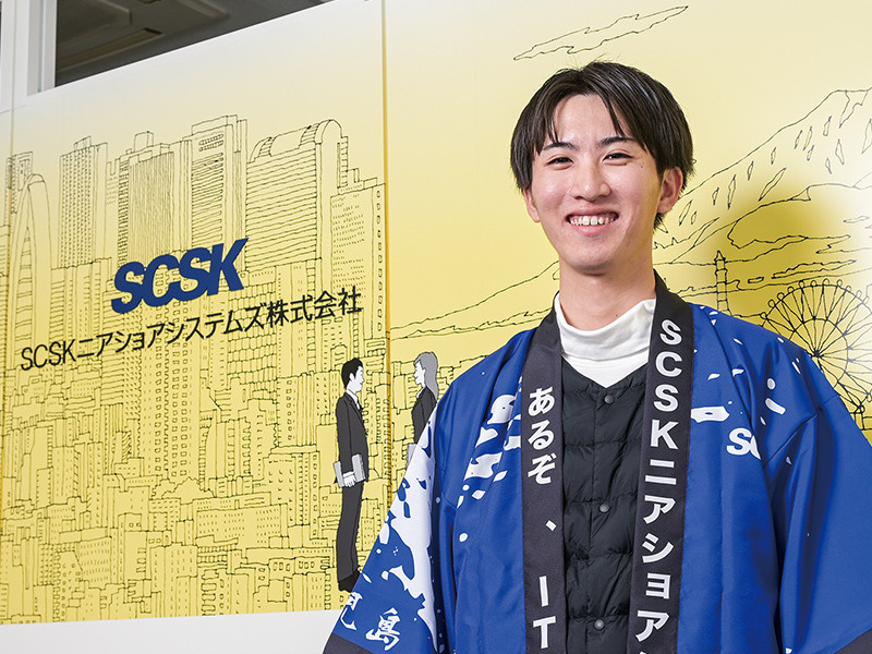 写真：青い法被を着て微笑んでいる男性。後ろの壁にはSCSKニアショアシステムズ株式会社のロゴマークと社名が掲げられている。