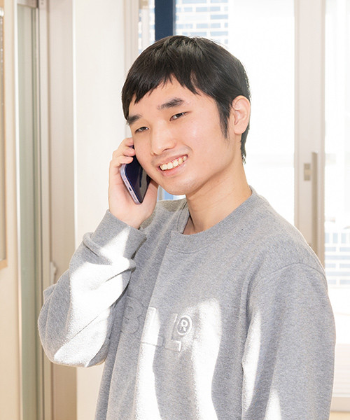 写真：グレーのスウェットを着た男子学生。スマートフォンで電話をかけている。