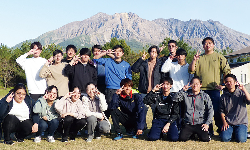 写真：全校レクリエーションの様子。18人の生徒が山を背に2列に並びピースサインをしている。