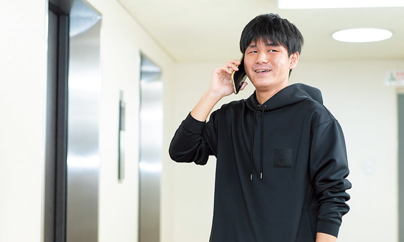 写真：黒いパーカーを着て、腕まくりをしている男子学生。スマートフォンで電話をかけている。