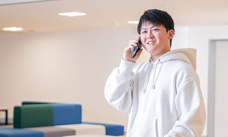 写真：白いパーカーを着た男子学生。スマートフォンで電話をかけている。