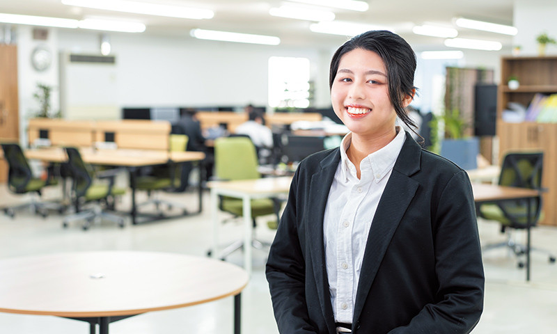 写真：シャツにジャケットを着た笑顔の女性。後ろには机や椅子が置かれたオフィスの風景が見える。