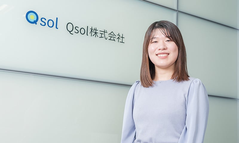 写真：薄灰色の服を着た笑顔の女性。後ろの壁にQsol株式会社のロゴマークと社名が掲げられている。