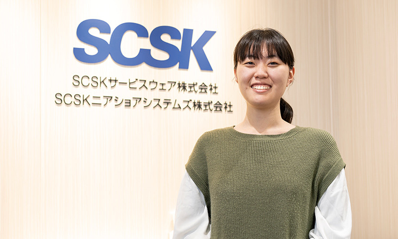 写真：白い服に緑のニットを着た笑顔の女性。後ろの壁にSCSKニアショアシステムズ株式会社のロゴマークなどが掲げられている。