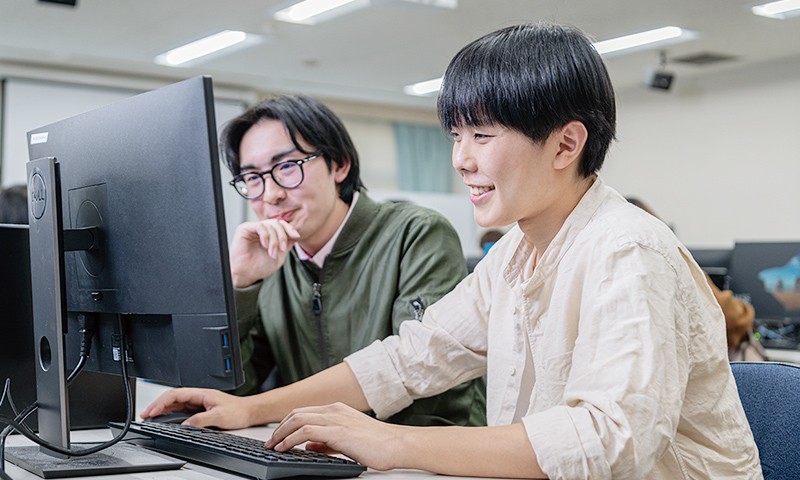 写真：黒木さんがデスクトップパソコンで作業をしている様子。隣では同級生の学生が一緒にモニターを見ている。