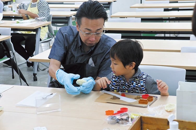 写真：エプロンと手袋をして、子どもといっしょに調理をしている小野先生。机の上には型抜きやヘラなどが置かれている。