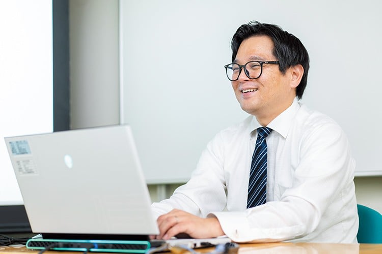 写真：シャツを着てネクタイを締めた、メガネを掛けている男性。ノートパソコンが置かれた机に向かって座り、微笑んでいる。