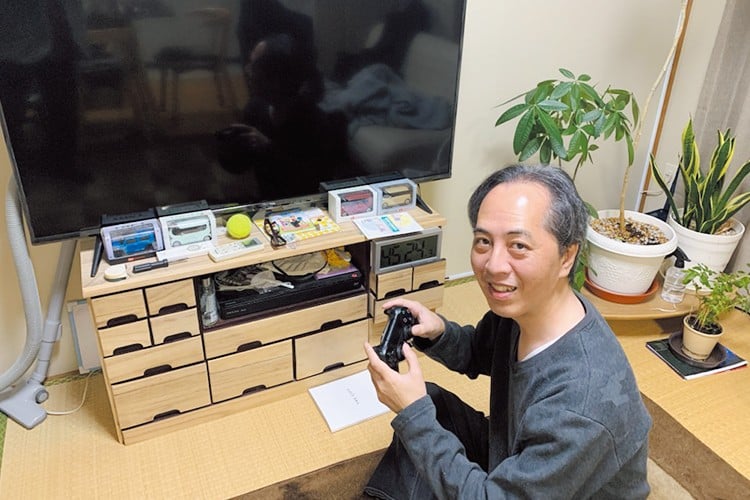 写真：テレビの前に座り、笑顔でゲームのコントローラーを持つ西先生