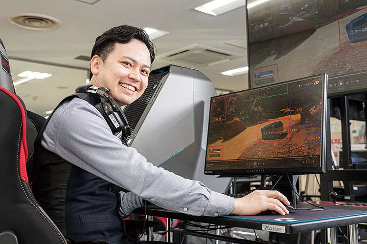 写真：机の前のゲーミングチェアに座り、微笑んでいる男性。机の上にはデスクトップパソコンとモニターが置かれている。