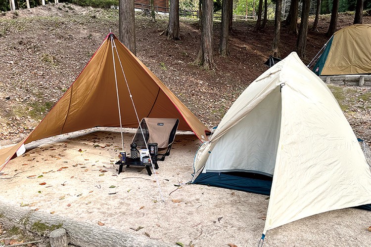 写真：木立のそばに茶色と白のテントが立てられている。左側の茶色のテントの中にはキャンプ用の机と椅子が置かれている。