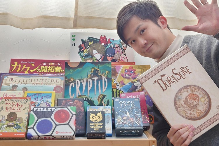 写真：ボードゲームのコレクションが机の上に並べられている。瀧野先生が机の隣でボードゲームを一つ持って微笑んでいる。