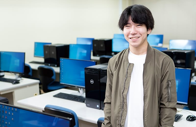 写真：白い服にモスグリーンの上着を着た男子学生。パソコンがたくさん置かれた教室の中に立ち、微笑んでいる。