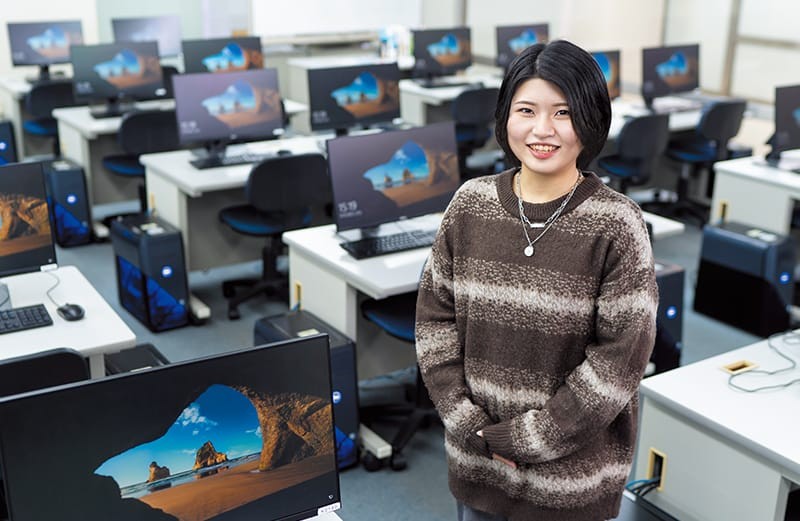 写真：茶色と白のストライプ柄のニットを着た女子学生。たくさんのパソコンが置かれた教室の中に立ち、微笑んでいる。