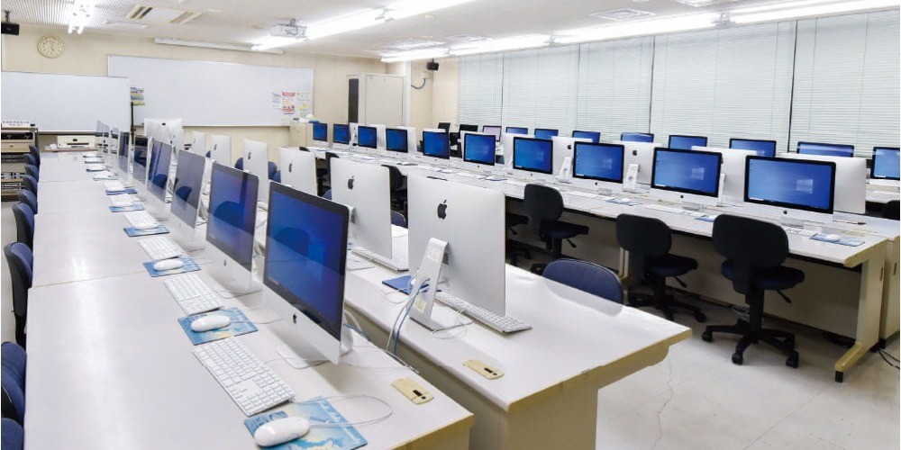 写真：左後ろ側から見た1F実習室。机がたくさん並んでおり、机の上にはiMacが置かれている。