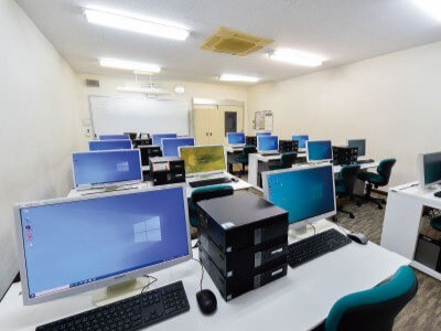 写真：左後ろ側から見た別館1F実習室。机がたくさん並んでおり、机の上にはWindowsのデスクトップパソコンが置かれている。