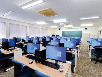 写真：右後ろ側から見た別館1F実習室。机がたくさん並んでおり、机の上にはWindowsのデスクトップパソコンが置かれている。