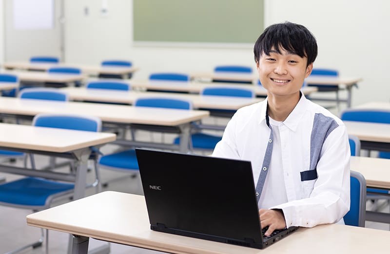 写真：シャツを着た男子学生。ノートパソコンが置かれた机に向かって座り、正面を向いて微笑んでいる。