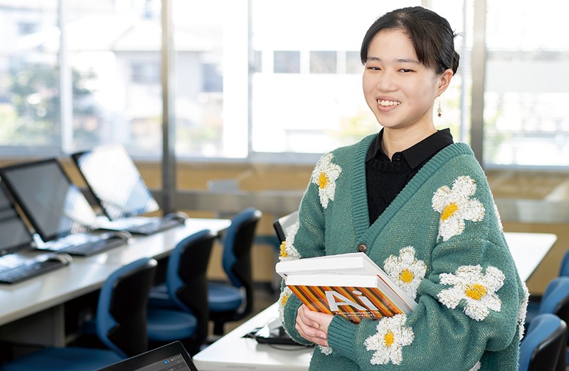 写真：黒いシャツに緑地に白い花柄のカーディガンを着た女子学生。両手でテキストを2冊持ち微笑んでいる。