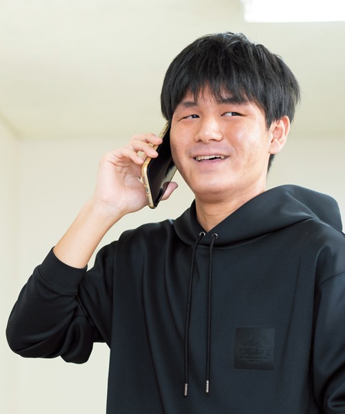 写真：黒いパーカーを着て、腕まくりをしている男子学生。スマートフォンで電話をかけている。