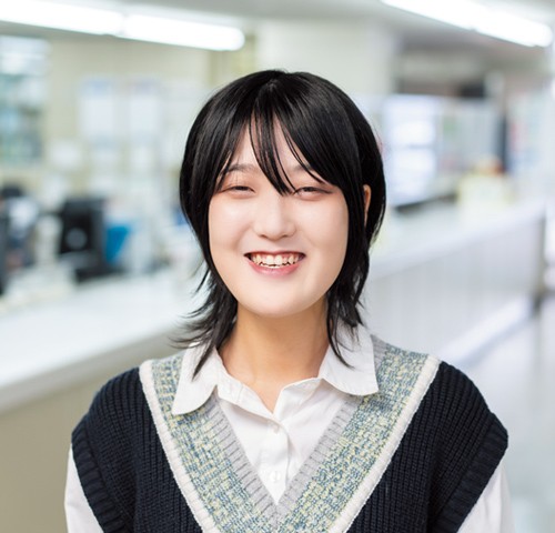 写真：白いシャツにVネックのニットを着た笑顔の女子学生