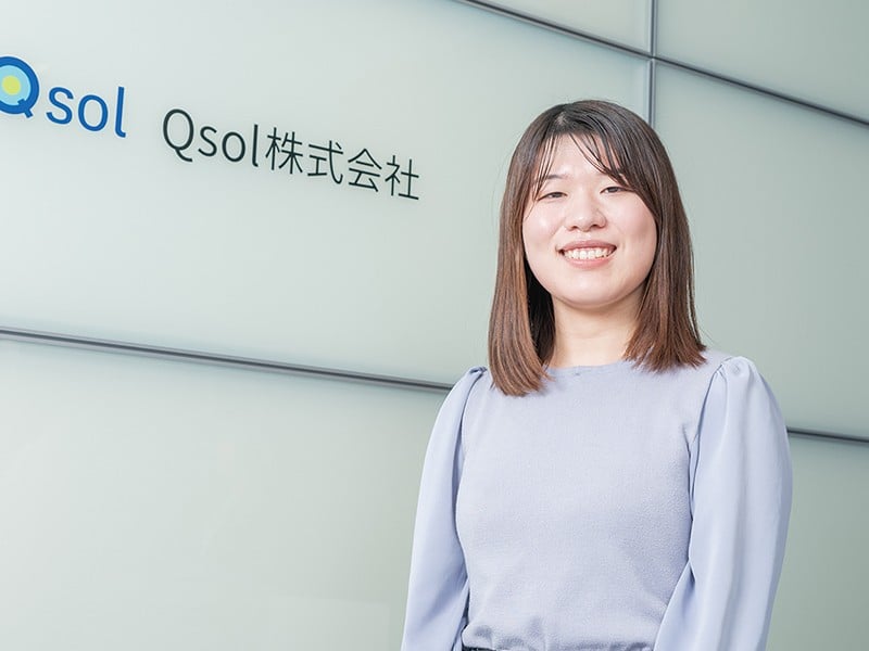 写真：薄灰色の服を着た笑顔の女性。後ろの壁にQsol株式会社のロゴマークと社名が掲げられている。