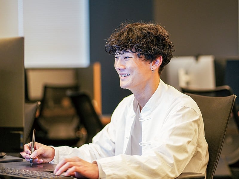 写真：白いチャイナシャツを着た笑顔の男性。パソコンが置かれた机に向かって座り、ペンタブレットを操作している。