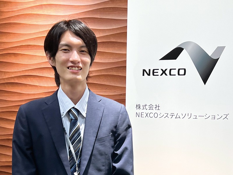 写真：青のスーツを着た笑顔の男性。後ろの壁には株式会社NEXCOシステムソリューションズのロゴマークと社名が掲げられている。