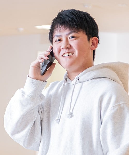 写真：白いパーカーを着た男子学生。スマートフォンで電話をかけている。