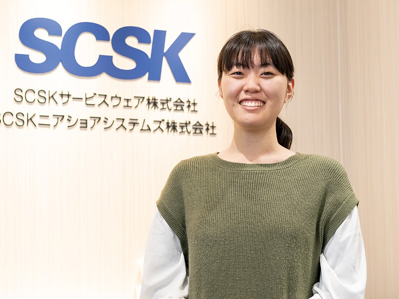 写真：白い服に緑のニットを着た笑顔の女性。後ろの壁にSCSKニアショアシステムズ株式会社のロゴマークなどが掲げられている。