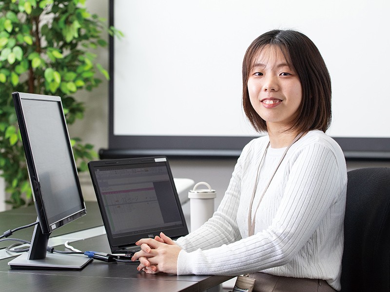 写真：白いニットを着て微笑んでいる女性。ノートパソコンやモニターが置かれた机の前に座っている。