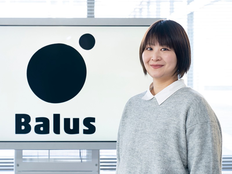 写真：白いシャツにグレーのニットを着た笑顔の女性。後ろのモニターにはBalusのロゴマークが表示されている。