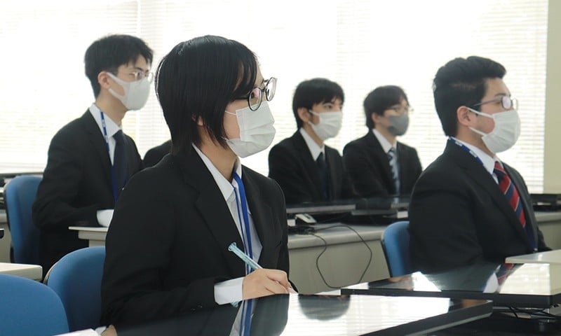 写真：企業説明会の様子。教室でスーツを着た学生が企業説明を聞いている。