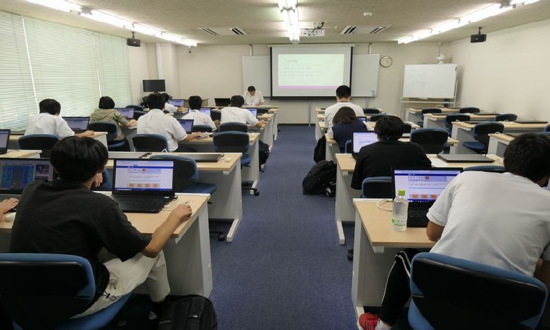 写真：プログラミングバトルの様子。学生5人がノートパソコンへ向かい作業をしている。