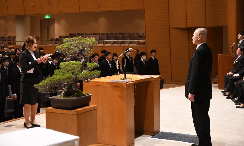 写真：入学式の様子。スーツを着た女子生徒がマイクの前に立ち、その少し後ろには他の生徒たちがそれぞれ椅子の前に立っている。