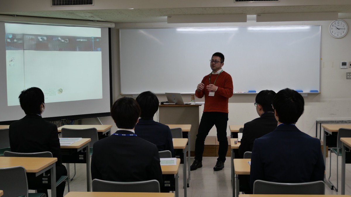 写真：企業説明会の様子。教室でスーツを着た学生が企業説明を聞いている。