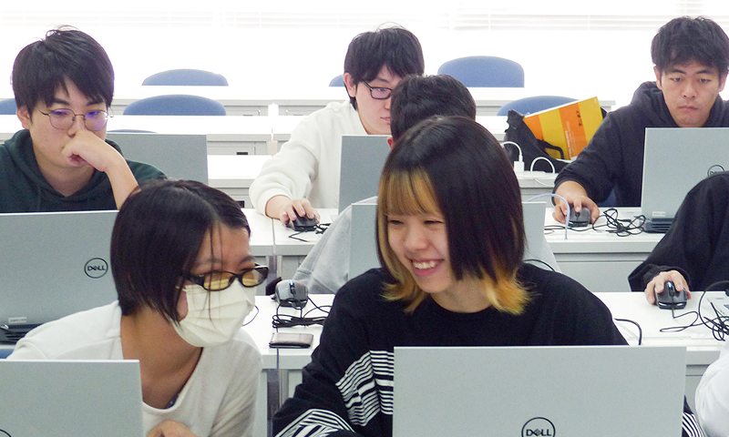 写真：プログラミングバトルの様子。数人の学生がノートパソコンへ向かい作業をしている。