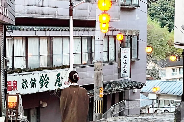 画像：温泉街の坂道を歩く茶色いコートを着た彼女の後ろ。周囲には旅館の看板や建物があり、街灯や提灯が並んでいる。