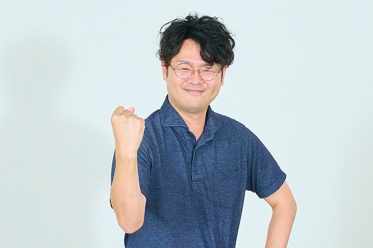 画像：メガネをかけた男性。彼は紺色のポロシャツを着ており、右手を握りしめてポーズを取っている。髪は少しカールした黒髪で、笑顔を見せている。