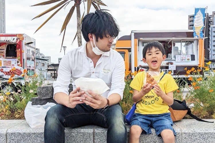画像：白いシャツの男性と黄色いTシャツの子どもが並んで座り、食べ物を手にしながら楽しい時間を過ごしている。