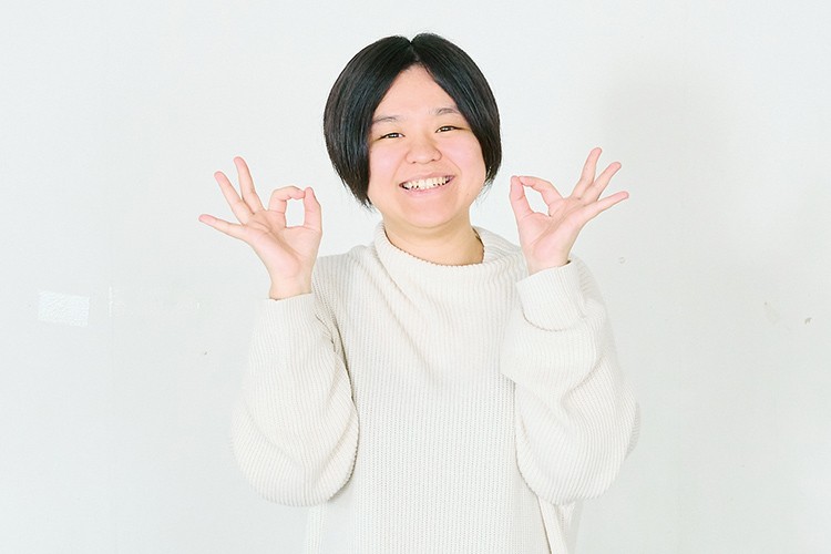 写真：白のニットセーターを着た笑顔の女性。両手で「OK」サインをしている。