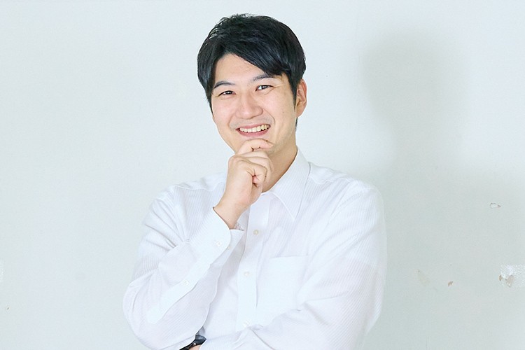 写真：白いシャツを着た笑顔の男性。片手をあごに添えてポーズをとっている。